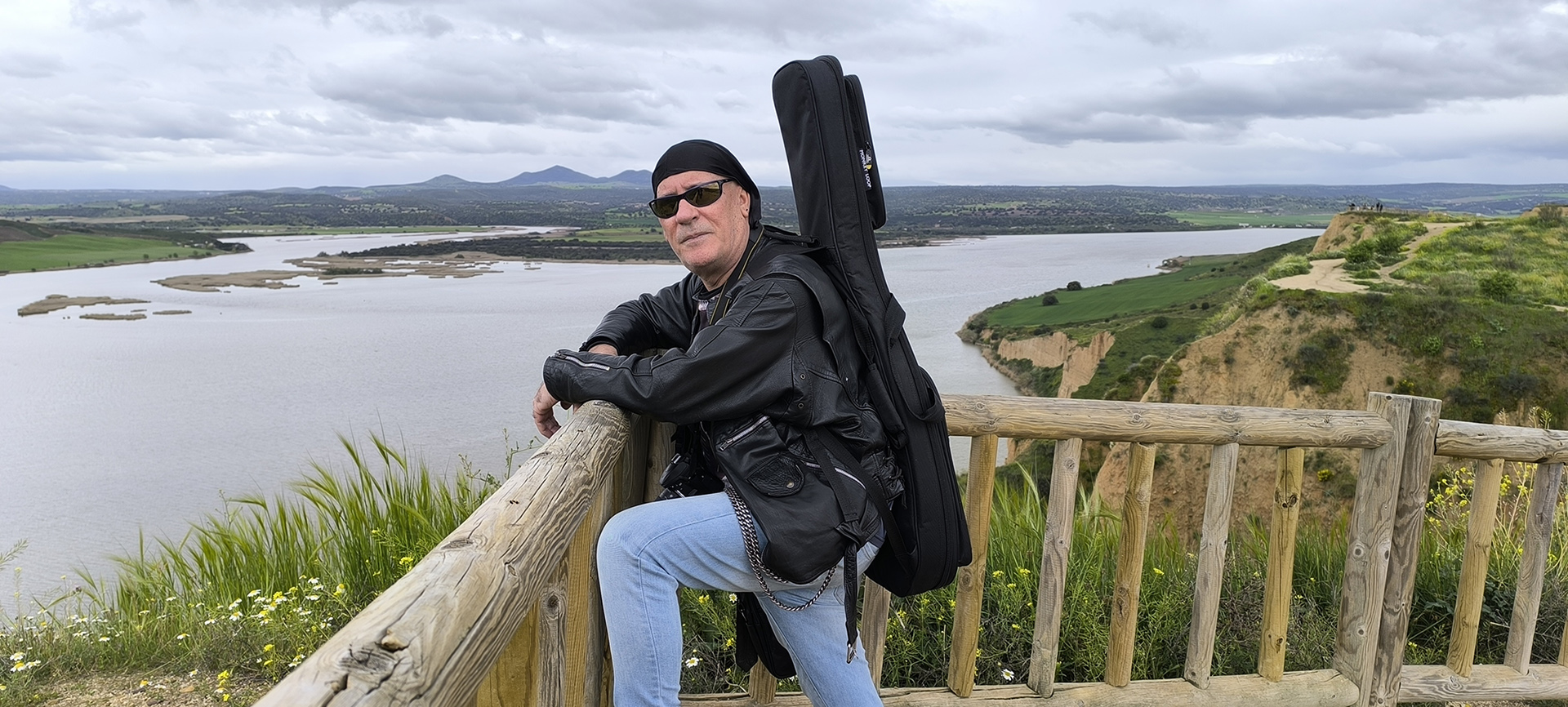 Jesús Valor con chaqueta de cuero y guitarra en la espalda, apoyado en una barandilla de madera.
